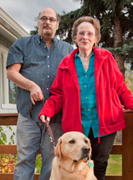 An elderly couple stand on the porch of their new home with their dog